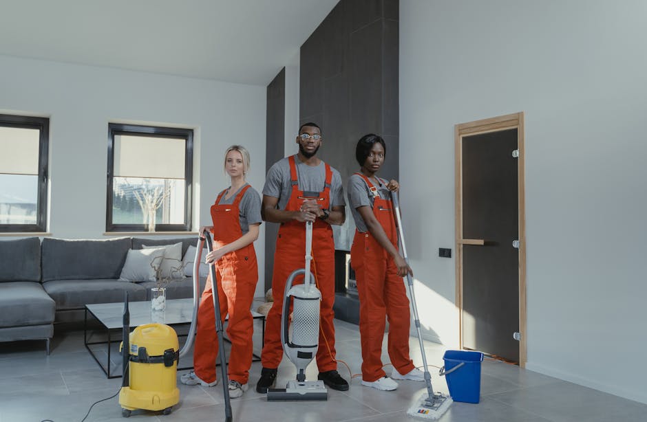 Three professional cleaners from Cleaners SW17 standing in a modern living room with a grey sofa, glass coffee table, and two large windows. They are dressed in grey T-shirts and red coveralls, holding cleaning tools such as vacuums, mops, and cloths. The room features light-colored walls, tiled flooring, and a black accent wall near a closed wooden door. The cleaners are positioned to demonstrate surface and deep cleaning tasks, emphasizing hygiene and maintenance, with sunlight illuminating the space to highlight its cleanliness and shine. The setting illustrates the high standards of cleaning service provided by [COMPANY_NAME] for residential spaces, aligning with the focus on avoiding hidden charges and ensuring thorough sanitisation.