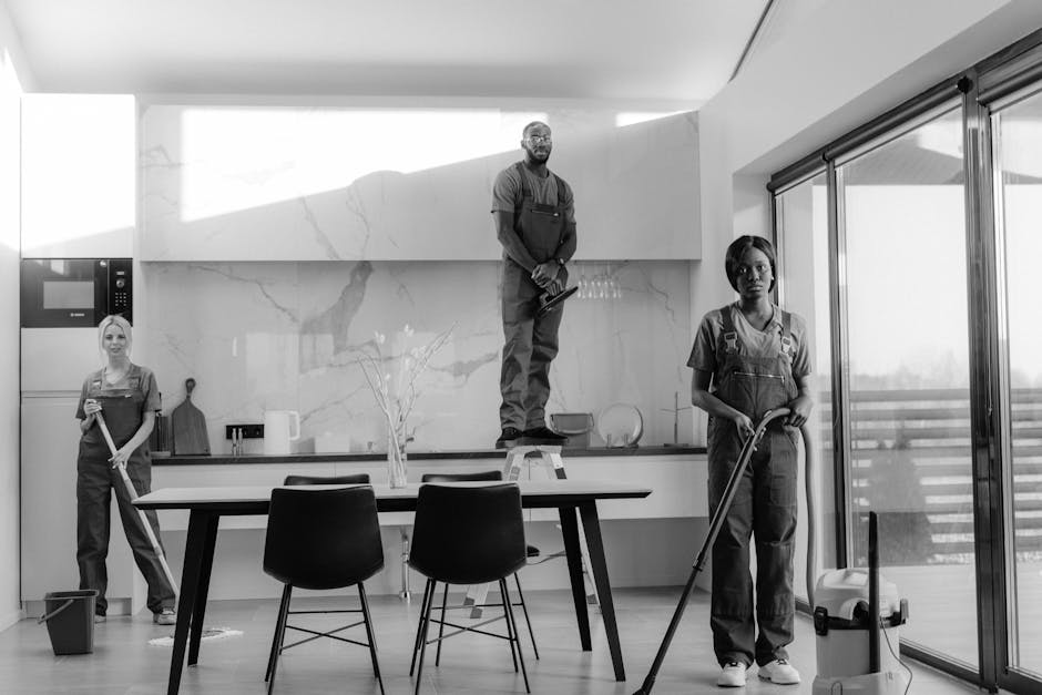 A team of three professional cleaners from Cleaners SW17 performing surface cleaning and deep cleaning in a modern living room. The room features a large L-shaped sofa with textured upholstery, a wooden coffee table, and two rectangular windows allowing natural light to illuminate the space. The floor is tiled with smooth, polished ceramic material, appearing clean and shiny. The cleaners are dressed in uniform with aprons, using vacuum cleaner, mop, and cloths to maintain hygiene and cleanliness. One cleaner is holding a vacuum hose, another is using a mop on the tiled floor, and the third has a cloth, suggesting routine sanitisation and dust removal. The room also includes a built-in wooden panel wall and a door in the background, with the overall scene showcasing an organised, well-maintained interior. This image emphasizes domestic cleaning processes that contribute to avoiding hidden charges, aligning with the purpose of the webpage from Cleaners SW17 about avoiding extra costs when booking cleaning services in Tooting, SW17.