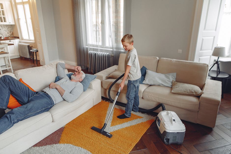A young boy using a vacuum cleaner to perform surface cleaning on a beige sofa in a well-lit living room. An elderly man is reclining on another sofa, resting with his eyes closed. The room features a wooden floor partially covered by a colorful orange and beige rug, with large windows allowing natural light to illuminate the space. The walls are painted light gray, and there are cushions on the sofas, along with a small side table with a lamp nearby. The vacuum cleaner is a modern, compact device, and the scene demonstrates domestic cleaning and maintenance in a high-traffic home environment, reflecting the focus of Tooting Broadway carpet cleaning guides for busy households. Cleaners SW17 offers professional cleaning services to ensure a hygienic, well-maintained living space.