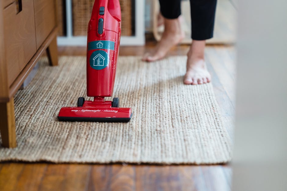A person using a red upright vacuum cleaner for surface cleaning on a beige textured carpet in a residential living room. The vacuum, which features a blue control button and a handle with icons indicating cleaning modes, is positioned near a wooden cabinet or sideboard. In the background, a person wearing black pants is barefoot, standing on the same carpet. Natural light illuminates the room, highlighting the cleanliness of the carpet, which appears well-maintained with an even texture. The wooden floor border is visible beneath the carpet, demonstrating the contrast between the polished hardwood and the clean, dust-free surface of the carpet. The scene emphasizes domestic deep carpet cleaning, showcasing the tools and conditions typical of high traffic home environments, aligning with the Tooting Broadway carpet cleaning guide for high traffic homes, SW17, offered by Cleaners SW17.