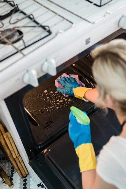 A person wearing blue rubber gloves, a yellow apron, and a white shirt is cleaning the interior of a black oven with a pink cloth, focusing on removing grease and stains from the oven surface. The oven is part of a white stove with visible burners on top, situated in a domestic kitchen with patterned black and white tile flooring. The lighting emphasizes the cleanliness and shine of the oven interior, reflecting a thorough surface cleaning and detailed sanitisation process, as promoted by Cleaners SW17 for same day oven cleaning services in Tooting SW17.